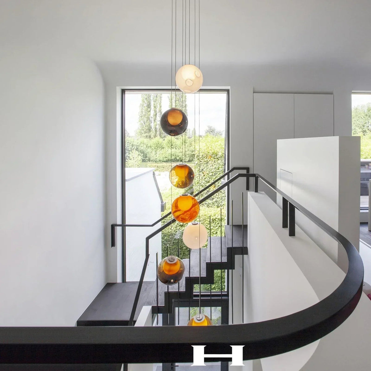 Chandelier with amber, smoky, and white glass globes descending through a bright stairwell with black railings and tall windows.