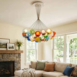 A colorful glass bubble chandelier hangs in a bright living room above a beige sectional sofa with decorative pillows, with a stone fireplace and large windows in the background.