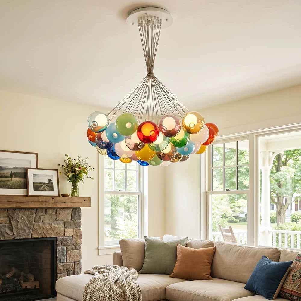 A colorful glass bubble chandelier hangs in a bright living room above a beige sectional sofa with decorative pillows, with a stone fireplace and large windows in the background.