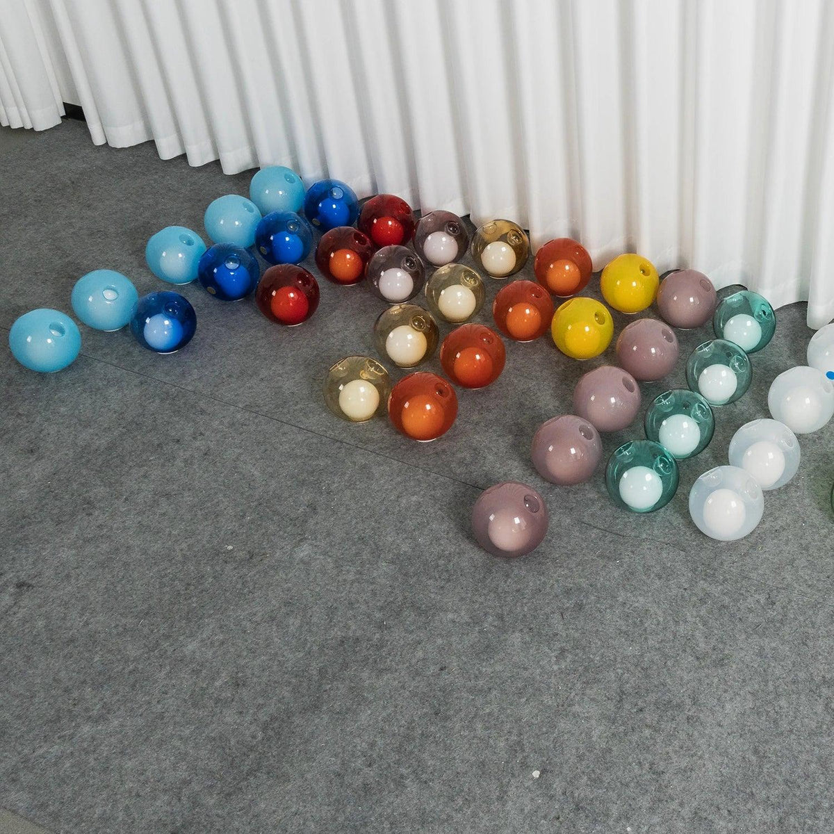 Chandelier glass globes in blue, red, amber, yellow, pink, and teal sit in rows on gray flooring beside white curtains.