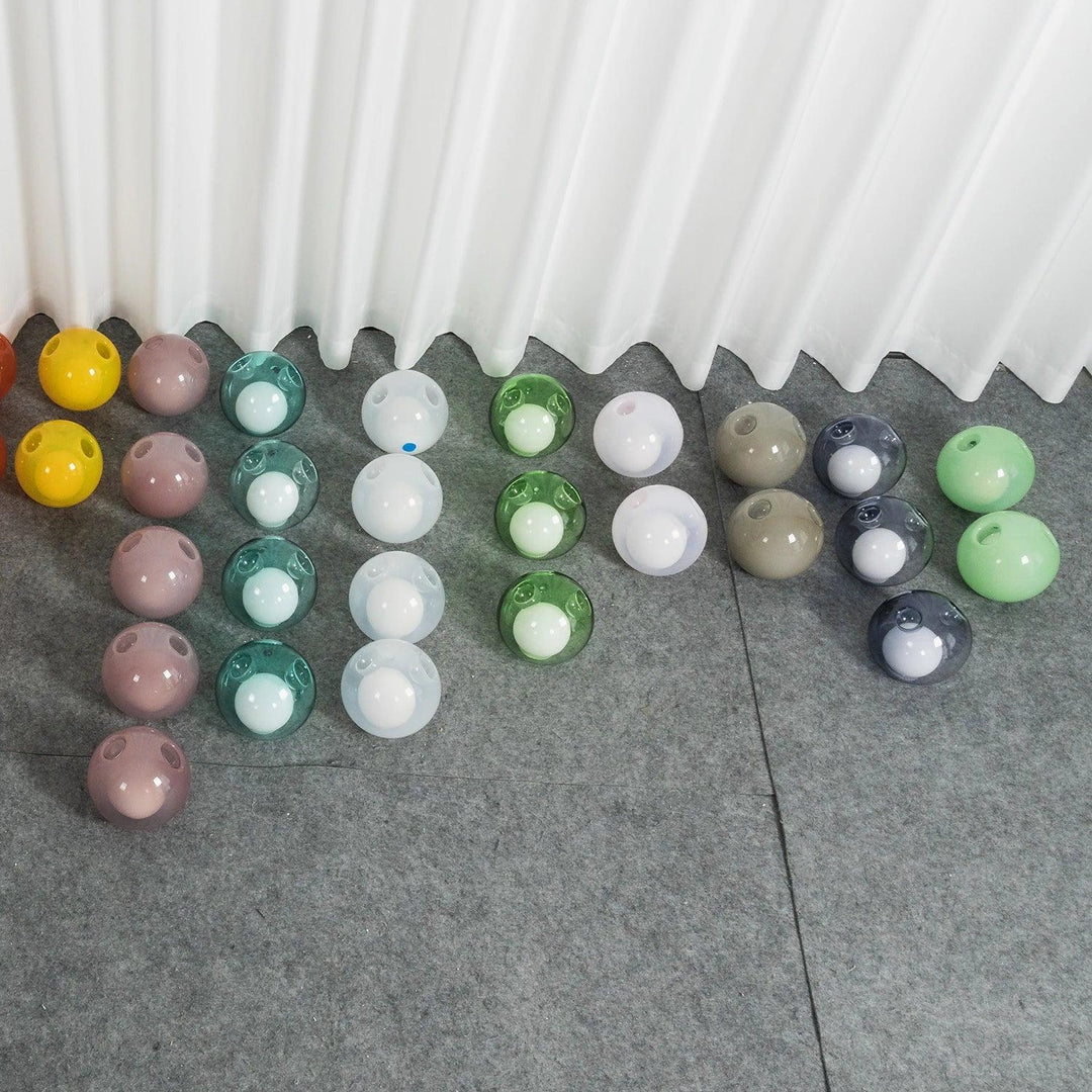 Glass globes for the chandelier in pink, teal, white, green, beige, and smoky purple sit on gray flooring beneath white curtains.