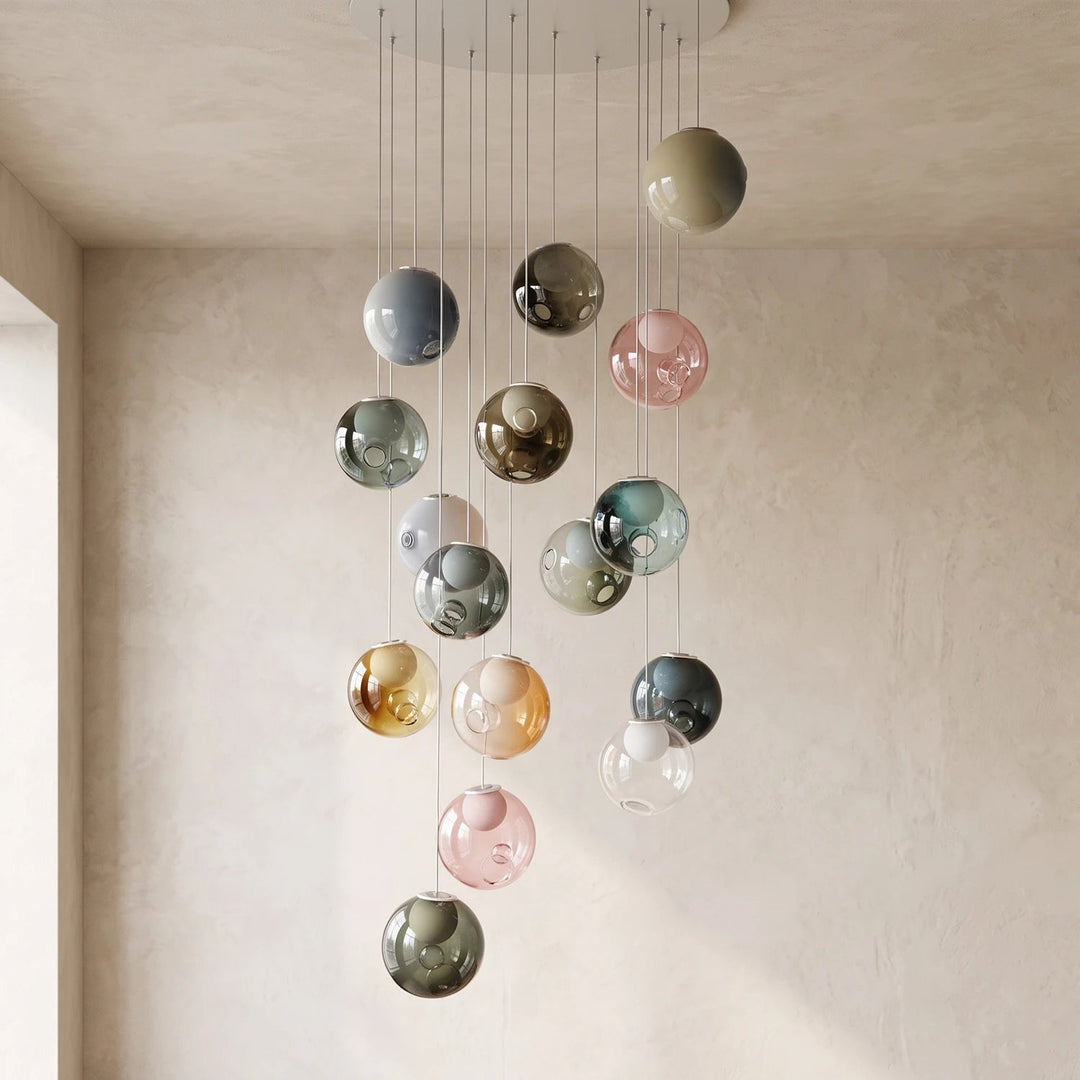 A modern multi-light chandelier featuring multiple hanging glass globes in assorted colors including pink, green, blue, gold, and gray, suspended from a circular ceiling mount against a textured beige wall.