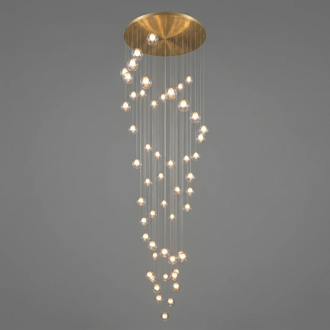 A modern multi-light chandelier with a brushed gold circular ceiling canopy and numerous clear glass spherical bulbs hanging in cascading vertical strands against a solid gray background.