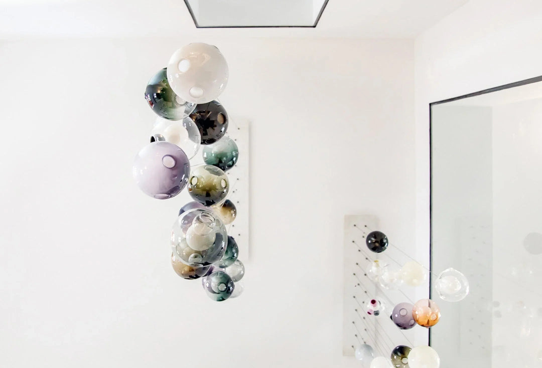 A chandelier with clustered glass globes in pale purple, teal, smoky and milky tones hangs in a white room beside a mirror.
