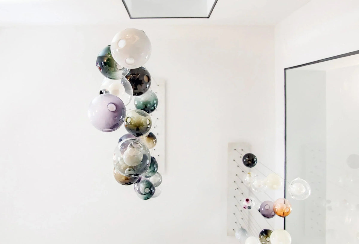 A chandelier with clustered glass globes in pale purple, teal, smoky and milky tones hangs in a white room beside a mirror.