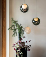 Chandelier with three glass globes in smoked grey and white hanging beside a green plant on a dark cabinet