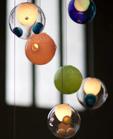 A chandelier close-up shows clear, orange, green, and blue glass globes with glowing inner lights against a windowed background.