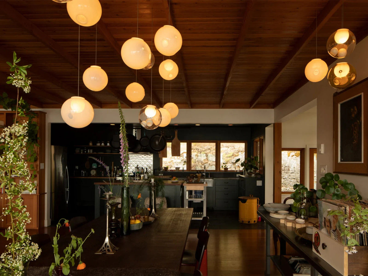 A chandelier with glowing milky and clear glass globes hangs beneath a wooden ceiling above a dining table adjoining a kitchen.