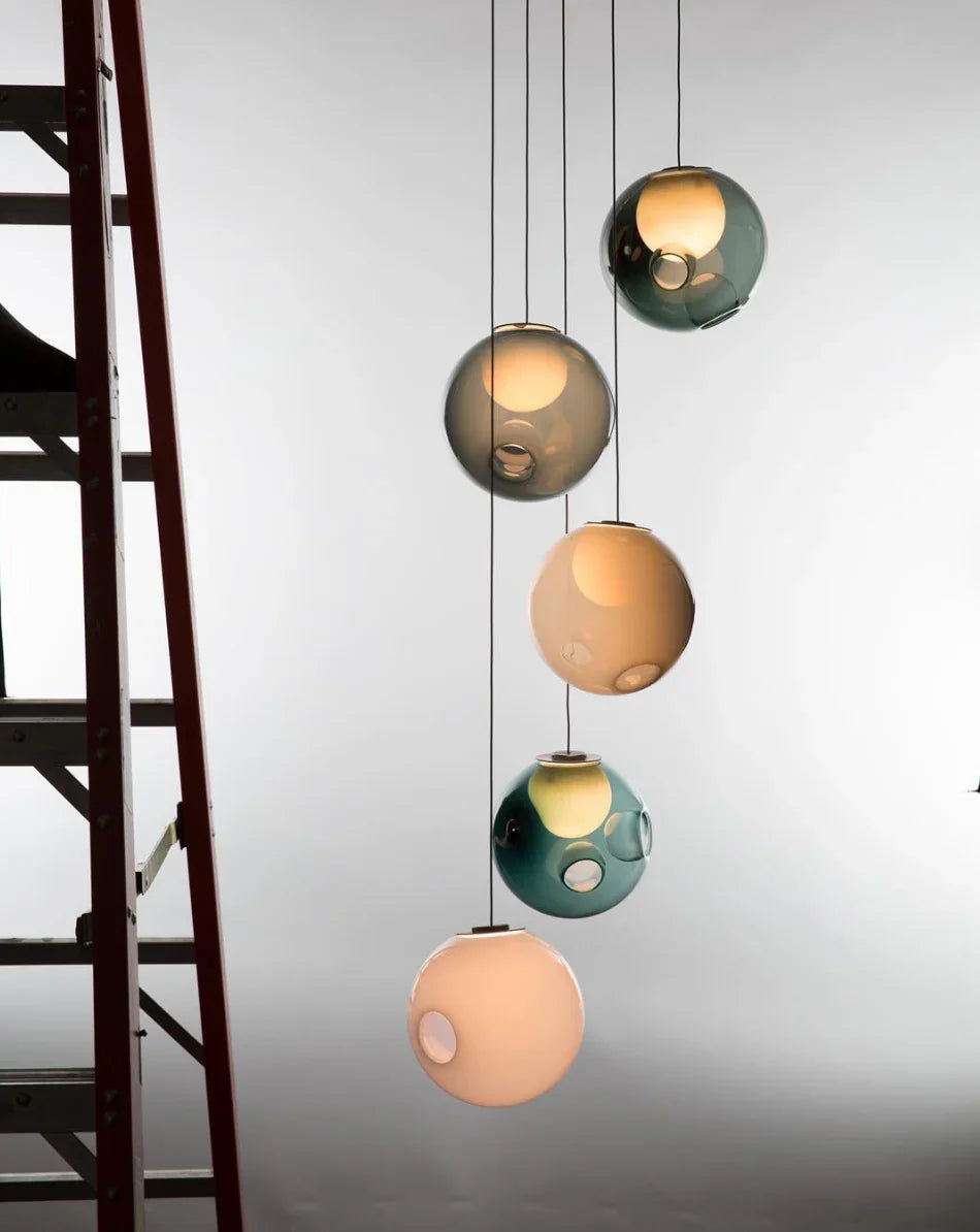 A five-globe chandelier with peach, teal, and smoky gray glass shades hangs beside a dark staircase against a pale wall.