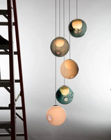 A five-globe chandelier with peach, teal, and smoky gray glass shades hangs beside a dark staircase against a pale wall.