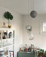 Three-globe glass chandelier with clear and smoky shades hangs at staggered heights above a home bar beside white shelving.