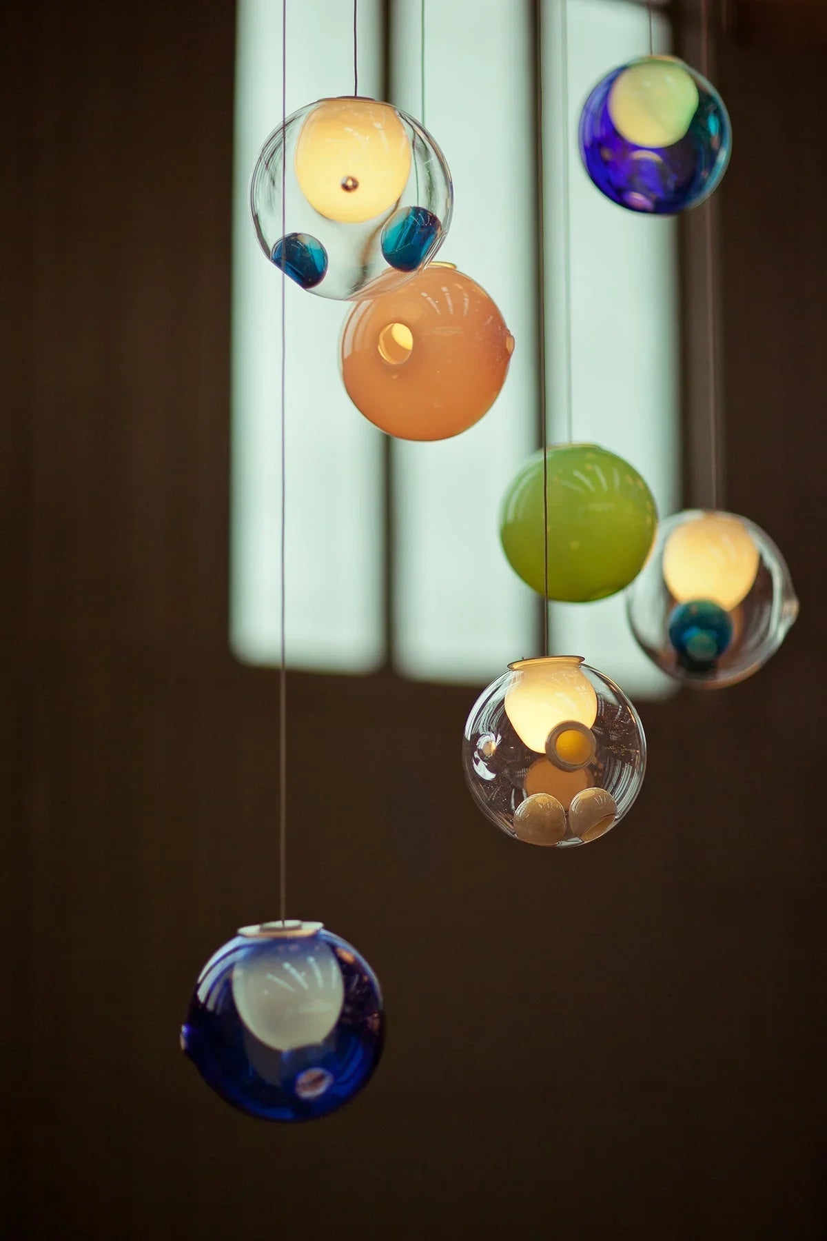 Multicolored glass globe chandelier with blue, green, amber, and clear shades hangs before a window in a dark interior.