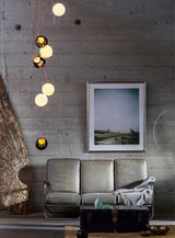 A modern living room scene featuring a three-seater sofa with a draped throw blanket, a dark trunk coffee table, a concrete wall, and a framed art print. A distinctive multi-light pendant chandelier with glowing white and amber glass spheres hangs on the left.