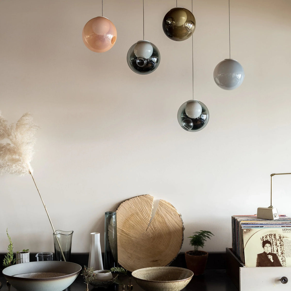 A chandelier with five pink, smoky and pale blue glass globes hangs above a sideboard displaying bowls, vases and records.