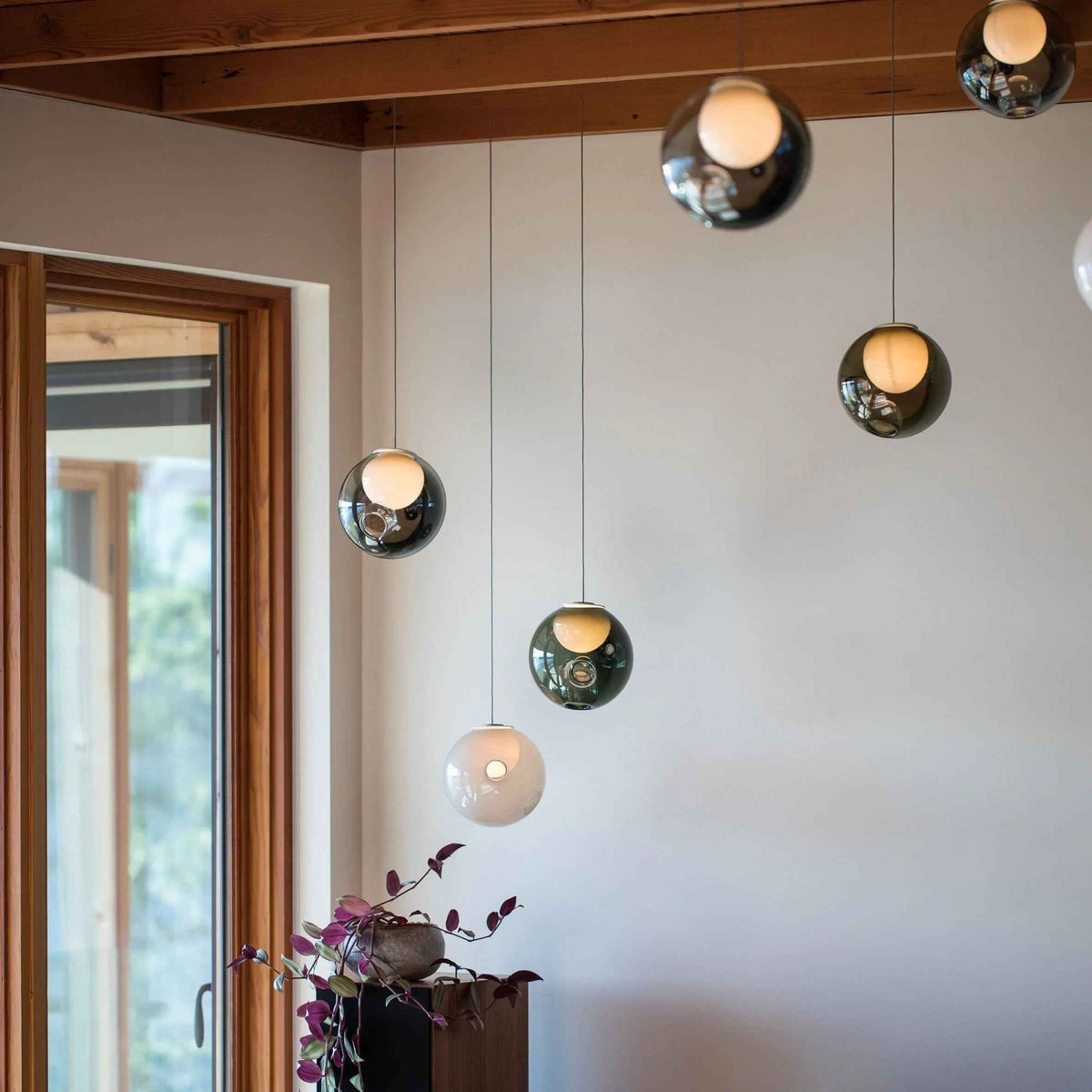 Chandelier with smoky and pale glass globes hanging beneath wooden ceiling beams beside a tall window and potted plant.