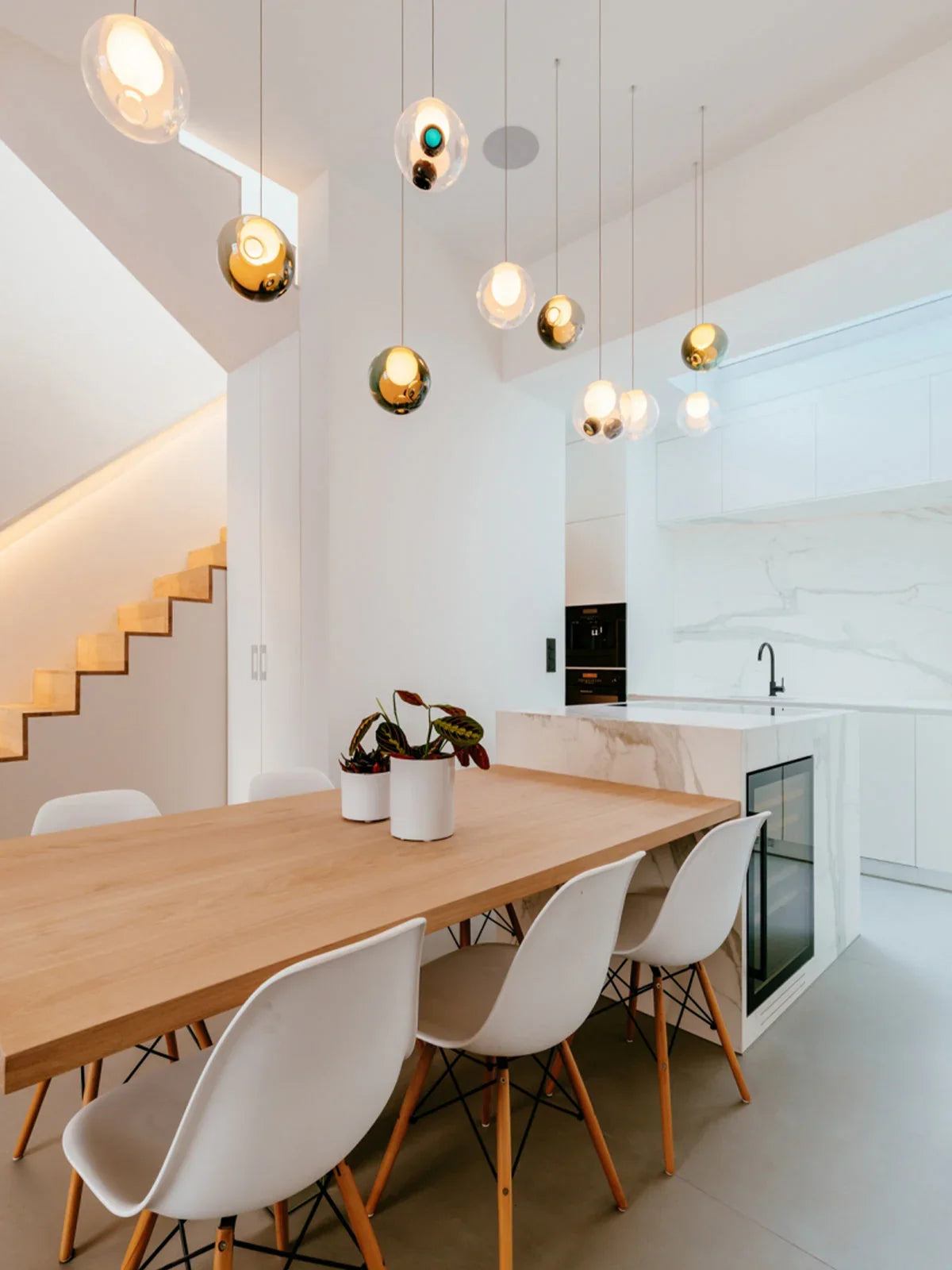 Glass globe chandelier lights hanging over a wooden dining table with white chairs in a bright kitchen with a staircase