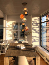 A modern multi-light pendant chandelier with multiple glass globe shades in orange, amber, and smoky tones, hanging over a rustic wooden dining table in a sunlit contemporary dining room.