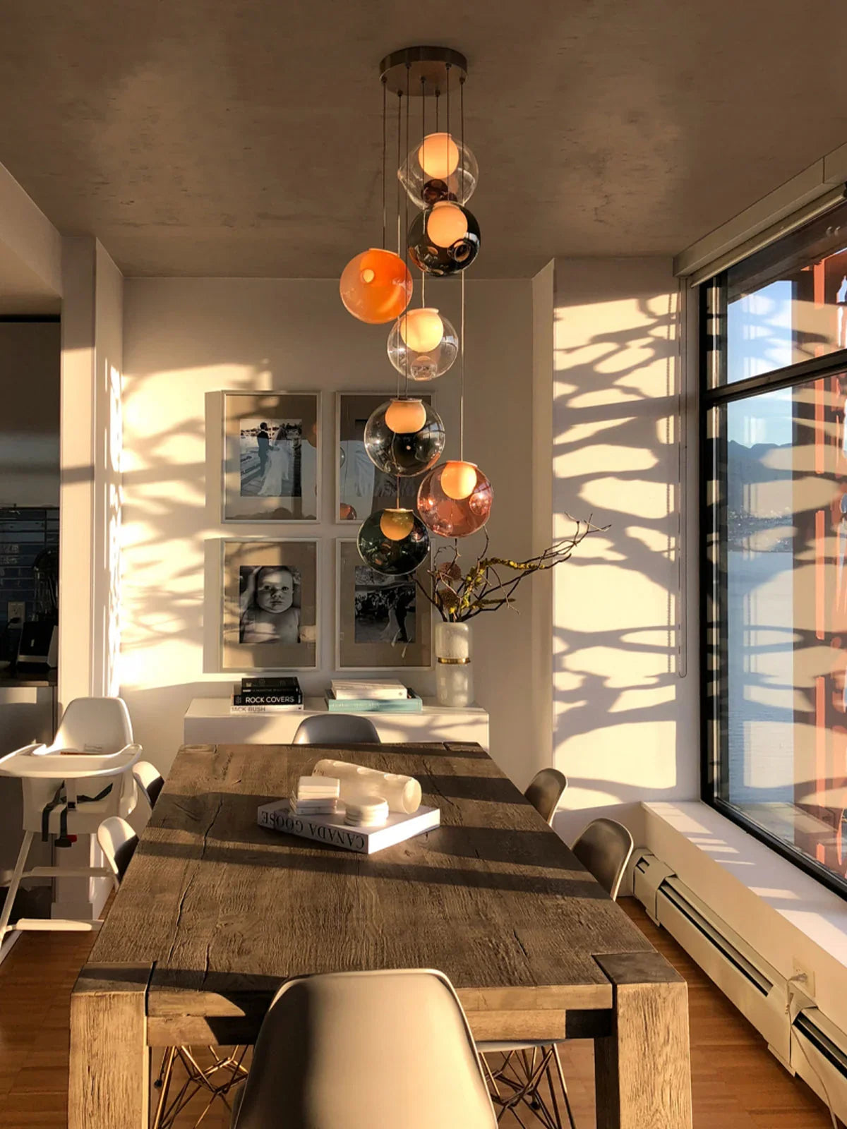 A modern multi-light pendant chandelier with multiple glass globe shades in orange, amber, and smoky tones, hanging over a rustic wooden dining table in a sunlit contemporary dining room.