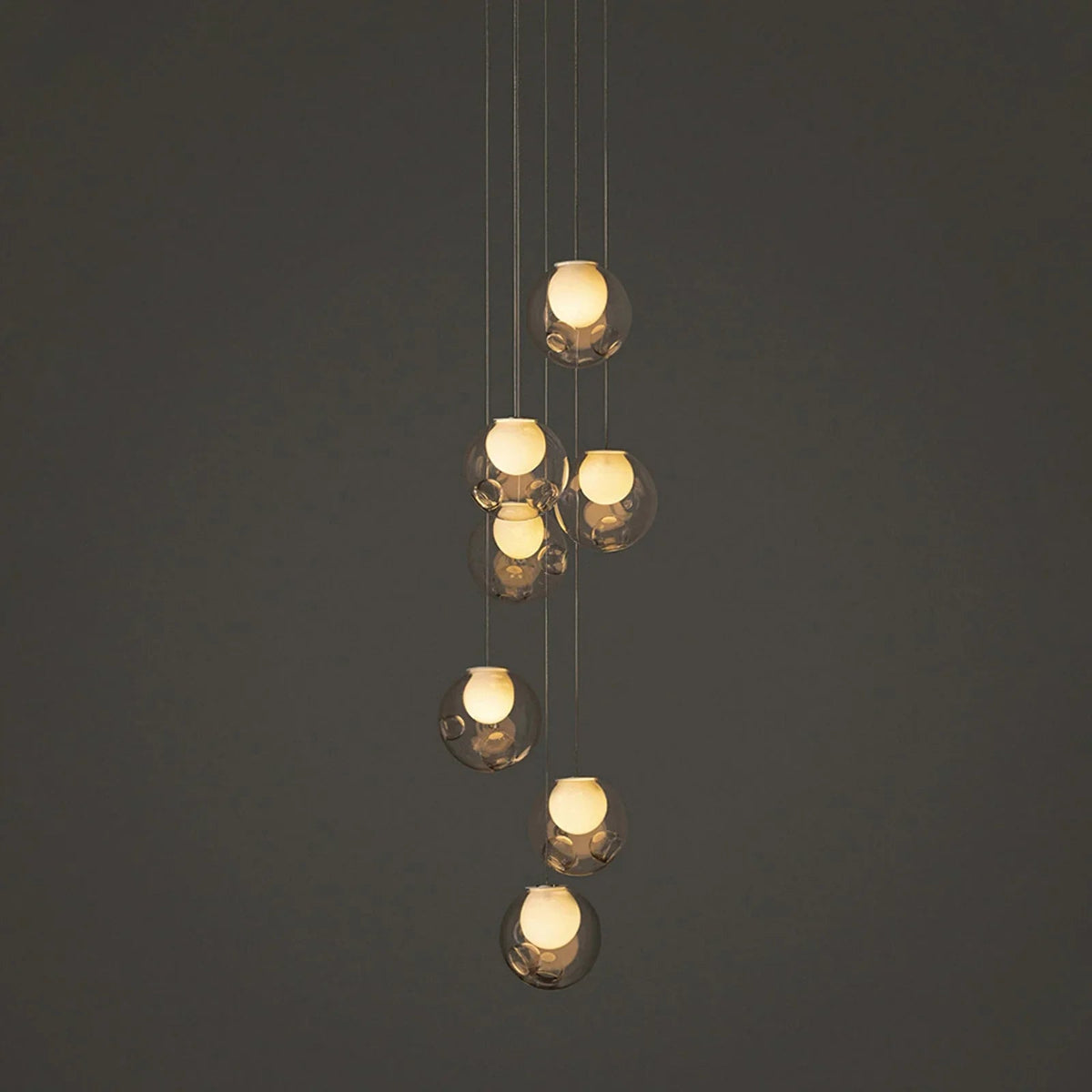 A modern multi-light pendant chandelier with seven clear glass bubble globes of varying sizes, suspended by thin cables against a dark gray background.