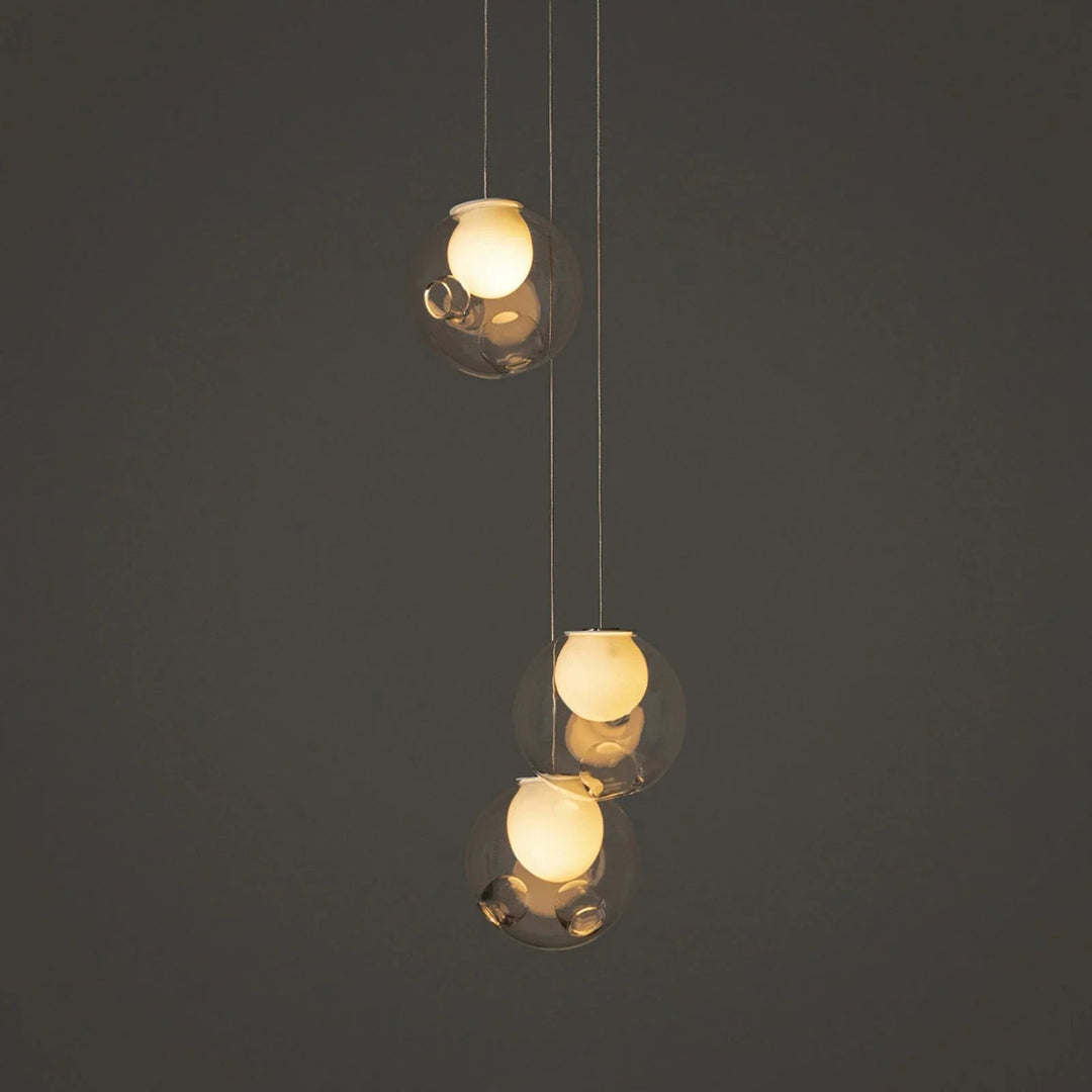 Three-light chandelier with clear glass globes and glowing cream-colored inner shades hangs at staggered heights against a dark gray background.