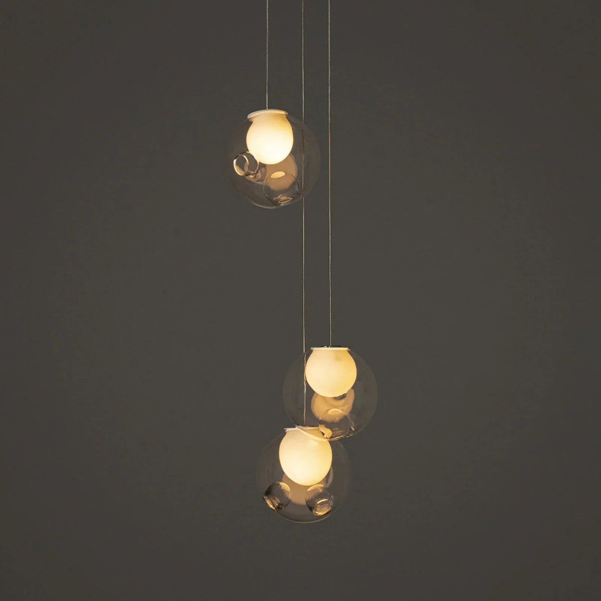 Three-light chandelier with clear glass globes and glowing cream-colored inner shades hangs at staggered heights against a dark gray background.