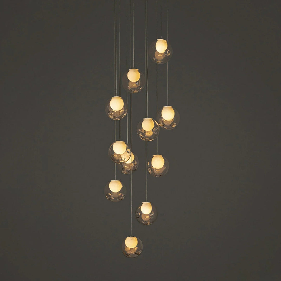 A modern multi-light pendant chandelier with clear faceted glass globes and warm glowing bulbs, suspended against a dark gray background.