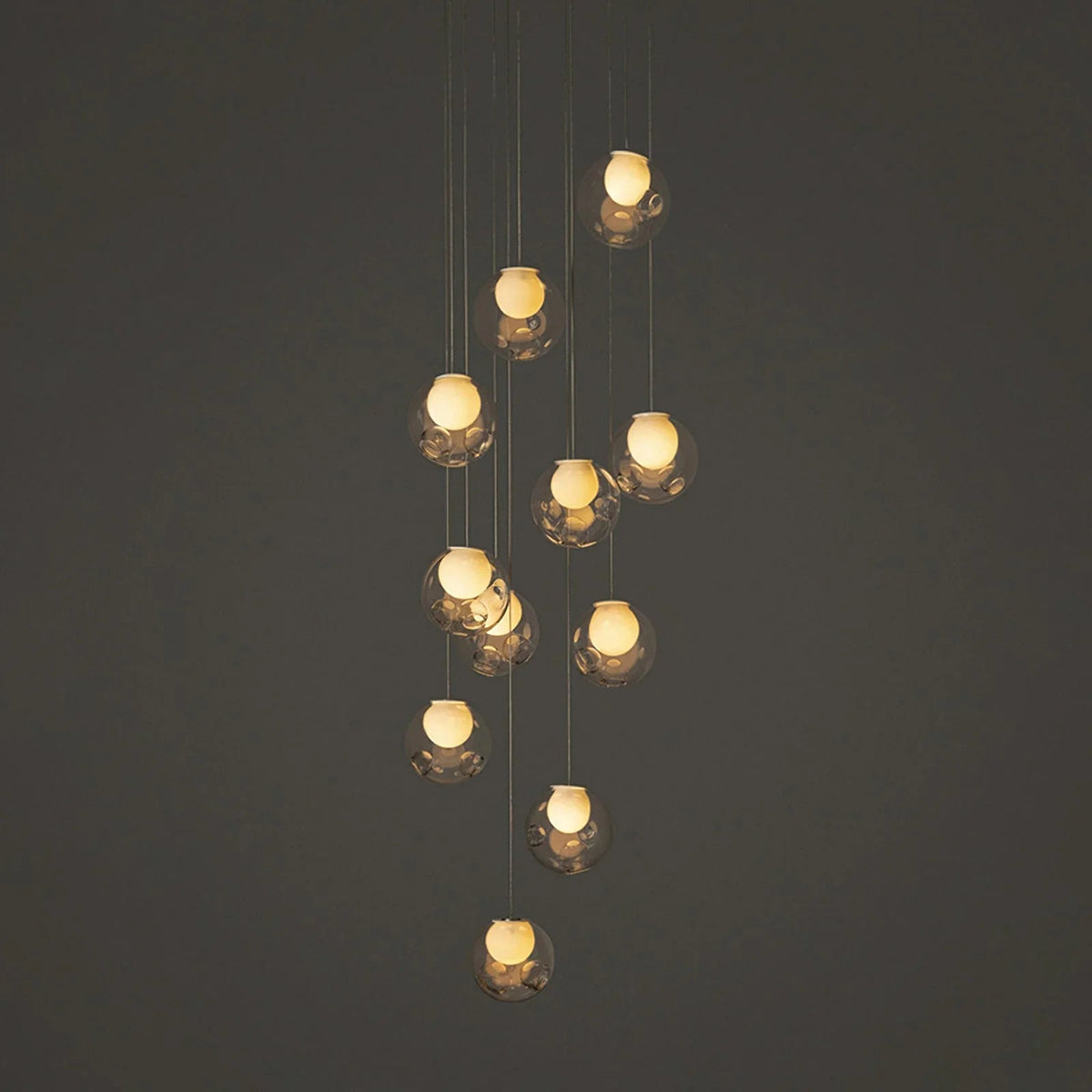 A modern multi-light pendant chandelier with clear faceted glass globes and warm glowing bulbs, suspended against a dark gray background.