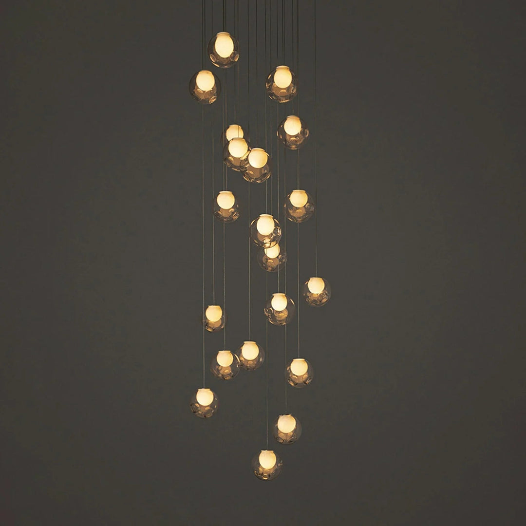 A modern multi-light linear chandelier with multiple warm glowing glass shades suspended at varying heights against a dark gray background.