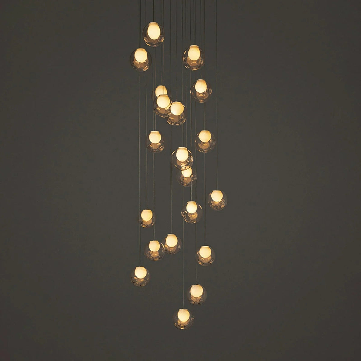 A modern multi-light linear chandelier with multiple warm glowing glass shades suspended at varying heights against a dark gray background.