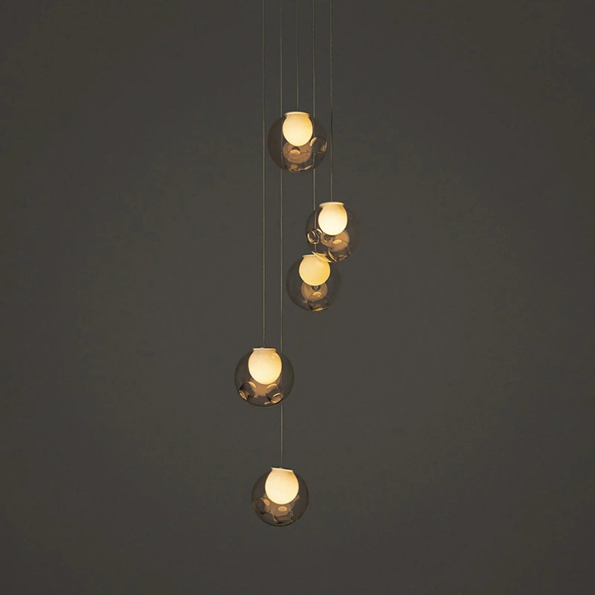 A modern multi-light pendant chandelier with five staggered, spherical glass globes suspended by thin cables against a dark gray background.