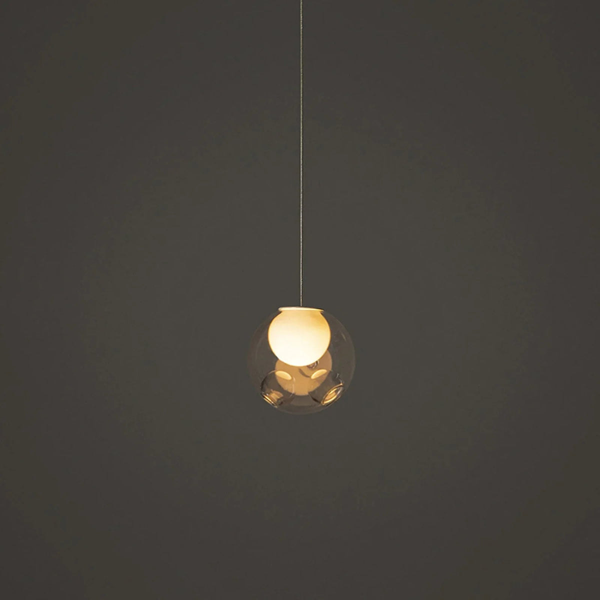 Single-light chandelier with a clear glass globe and glowing cream-colored inner shade hangs against a dark gray background.