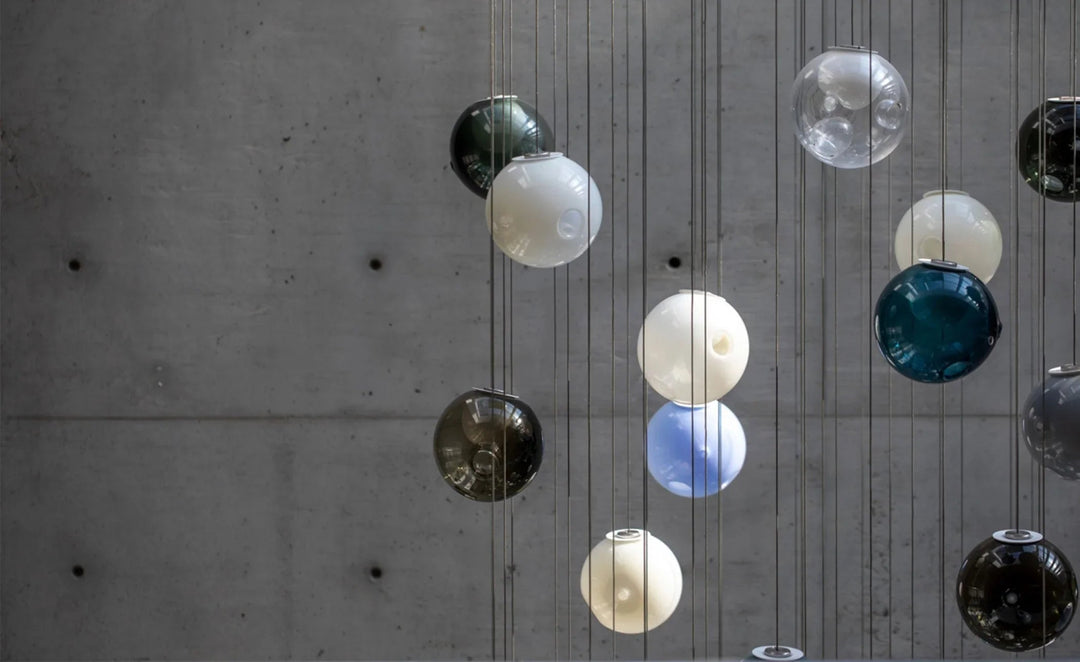 A chandelier with white, clear, blue, teal, and smoky glass globes hangs on thin cords against a gray concrete wall.