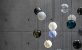 A chandelier with white, clear, blue, teal, and smoky glass globes hangs on thin cords against a gray concrete wall.