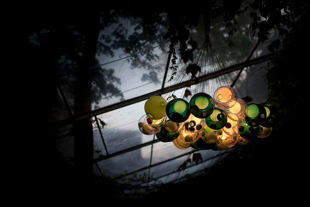 A chandelier with clustered green, yellow, and amber glass globes hangs beneath a glass roof framed by dark foliage.
