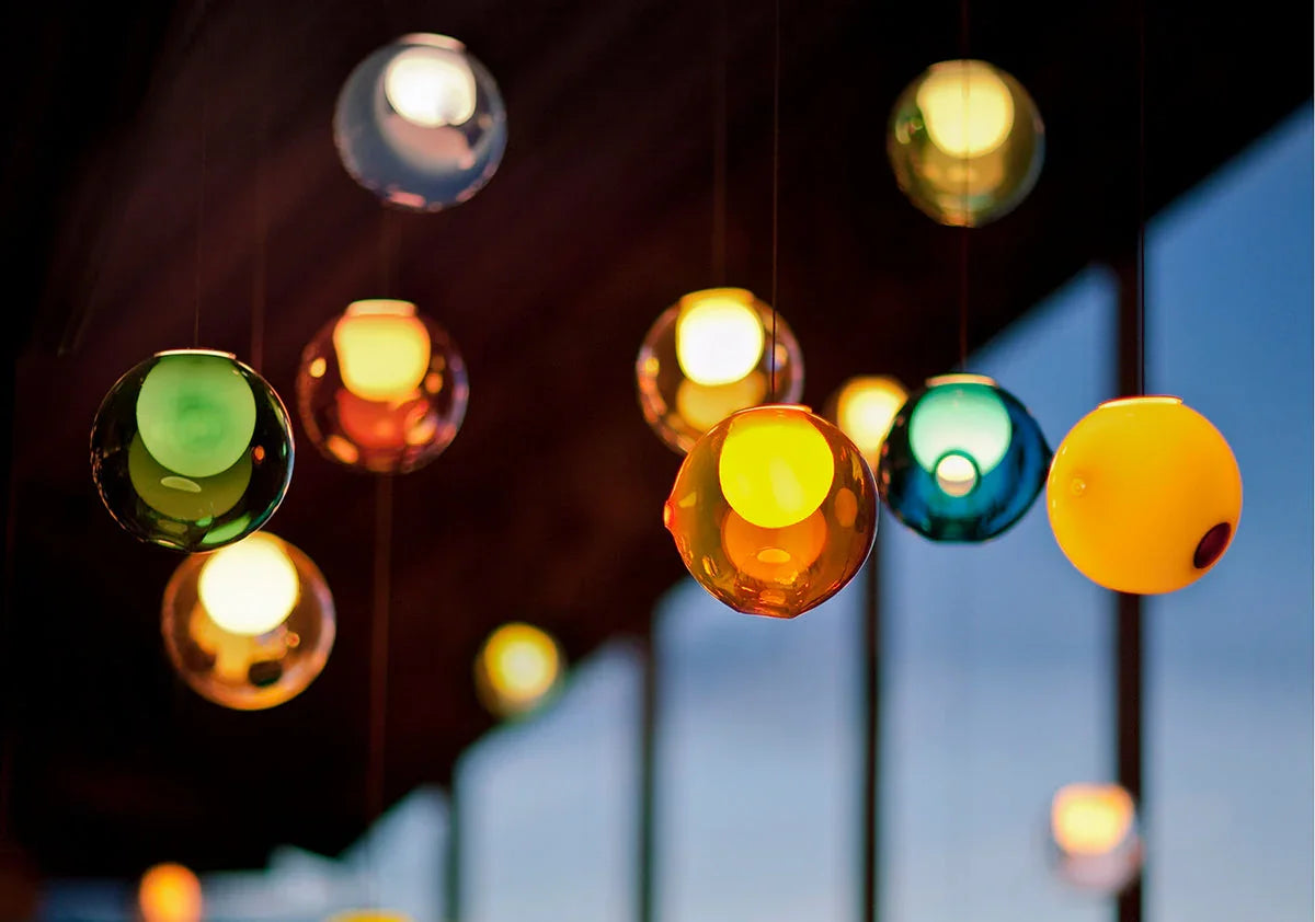A chandelier with illuminated green, amber, blue, and yellow glass globes hangs at varying heights beside tall windows.