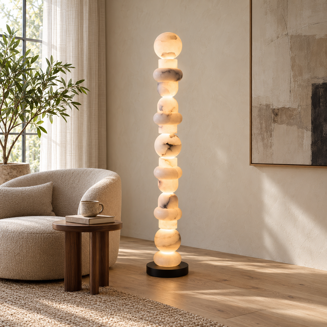 A modern floor lamp made of stacked glowing stone spheres on a black base, shown in a styled living room next to a beige armchair and small wooden side table.