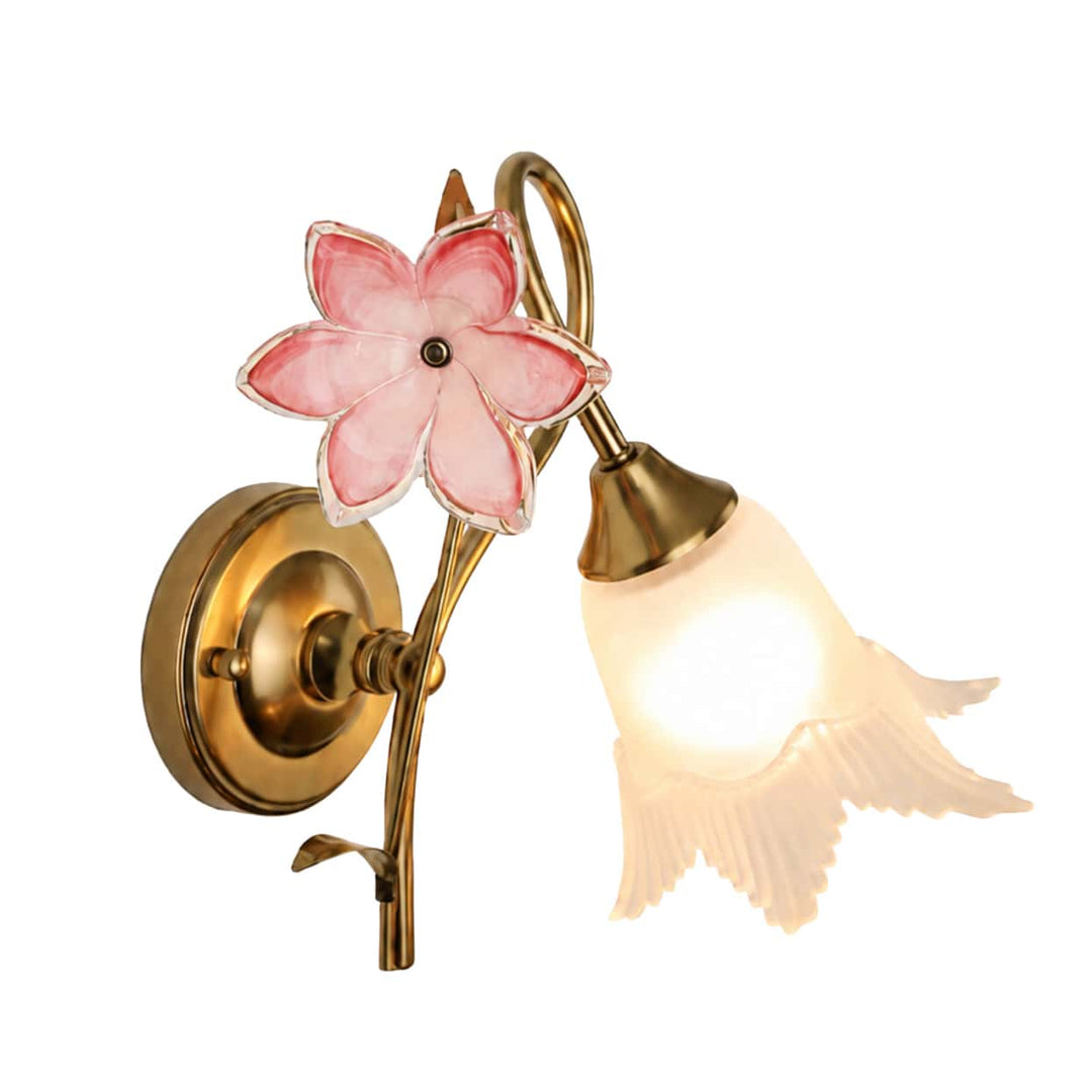 Gold-toned wall sconce with a pink flower accent and glowing cream bell-shaped shade against a white background.
