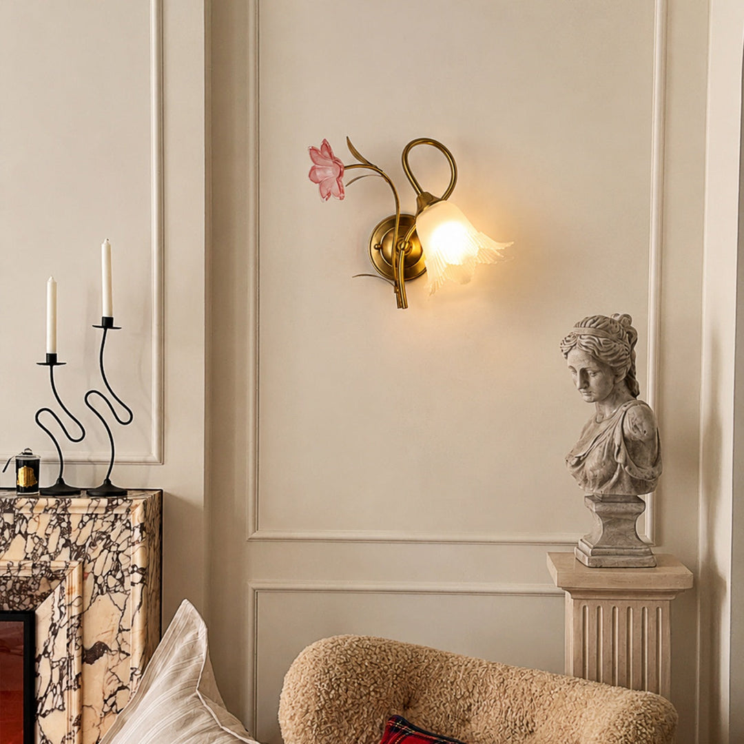 A decorative wall sconce with a gold-toned metal frame, a pink flower accent, and a glowing white globe bulb, mounted on a beige paneled wall in a styled interior setting.