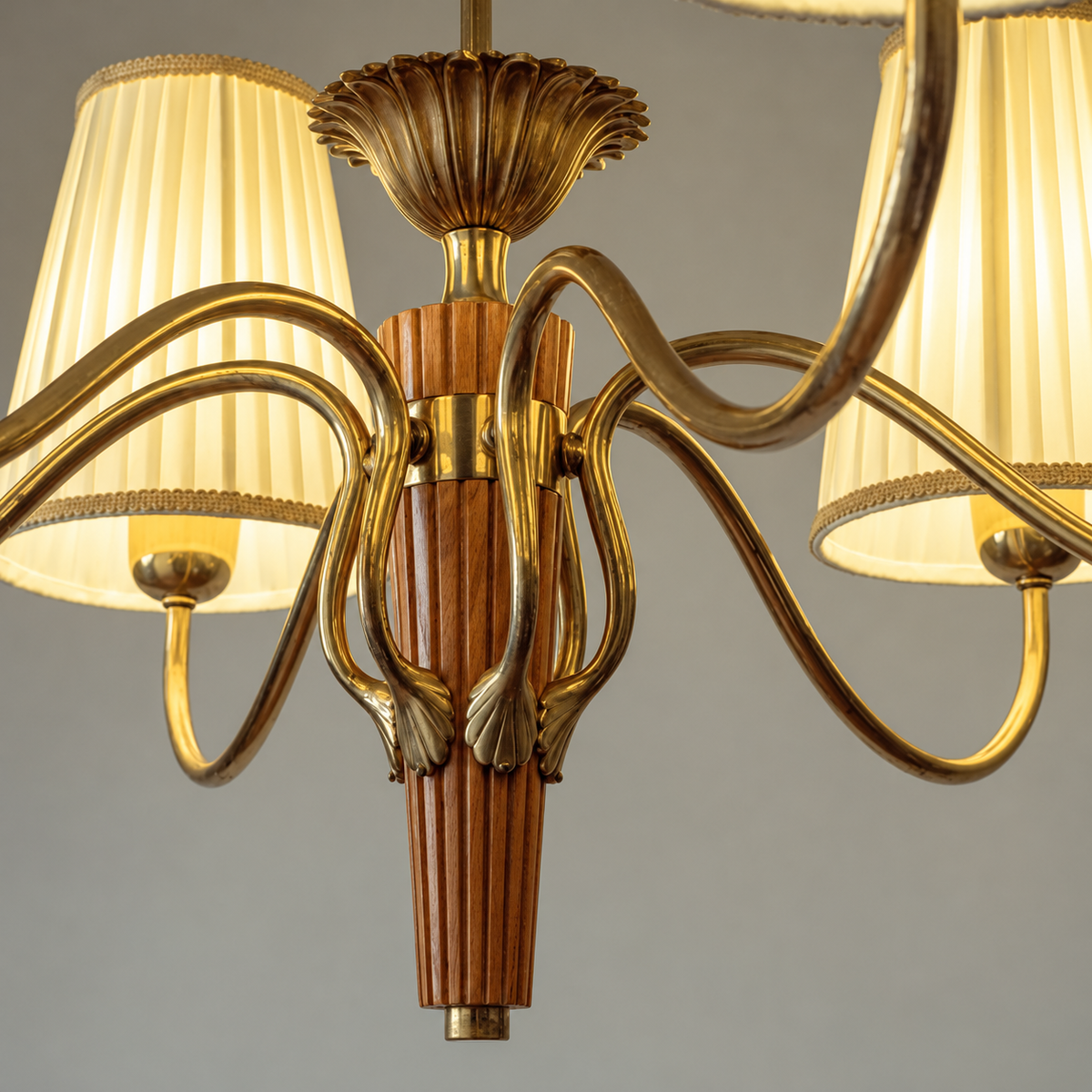 A close-up of a multi-arm chandelier with a polished gold brass finish, featuring curved arms, a fluted central column with warm wood accents, and pleated cream-colored fabric shades.