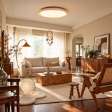 A modern circular flush-mount ceiling light with a warm wood trim and glowing frosted glass, installed in a beautifully styled, sunlit living room with beige walls, a wooden coffee table, and a rattan armchair.
