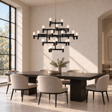 A modern multi-light chandelier with a matte black frame and warm glowing bulbs hangs over a dark wood dining table and six upholstered chairs in a bright, sunlit dining room.