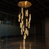 A modern multi-tier chandelier with cylindrical glass crystal rods emitting warm light, suspended from a circular gold ceiling canopy in a dark, contemporary indoor lobby.