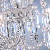Close-up of a crystal chandelier detail with clear faceted glass drops and prisms on a silver-toned metal frame, set against a softly blurred background of similar hanging crystal elements.