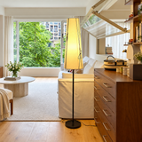 A modern floor lamp with a tall rectangular beige shade and black stand is positioned in a bright, contemporary living room next to a white sofa and wooden furniture.