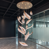 A modern multi-light feather chandelier with clear crystal-like leaves and warm copper-toned metal accents, suspended from a circular ceiling fixture in a contemporary office lobby with glass walls and a dark ceiling.