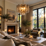A beautifully styled living room featuring a large, textured glass chandelier hanging above a fireplace. The room is filled with warm natural light from large black-framed windows, decorated with potted plants, a cozy armchair, and a coffee table with books and candles.