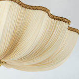 Close-up of a ceiling light with pleated cream fabric and gold-toned braided trim along its scalloped edge against a pale background.