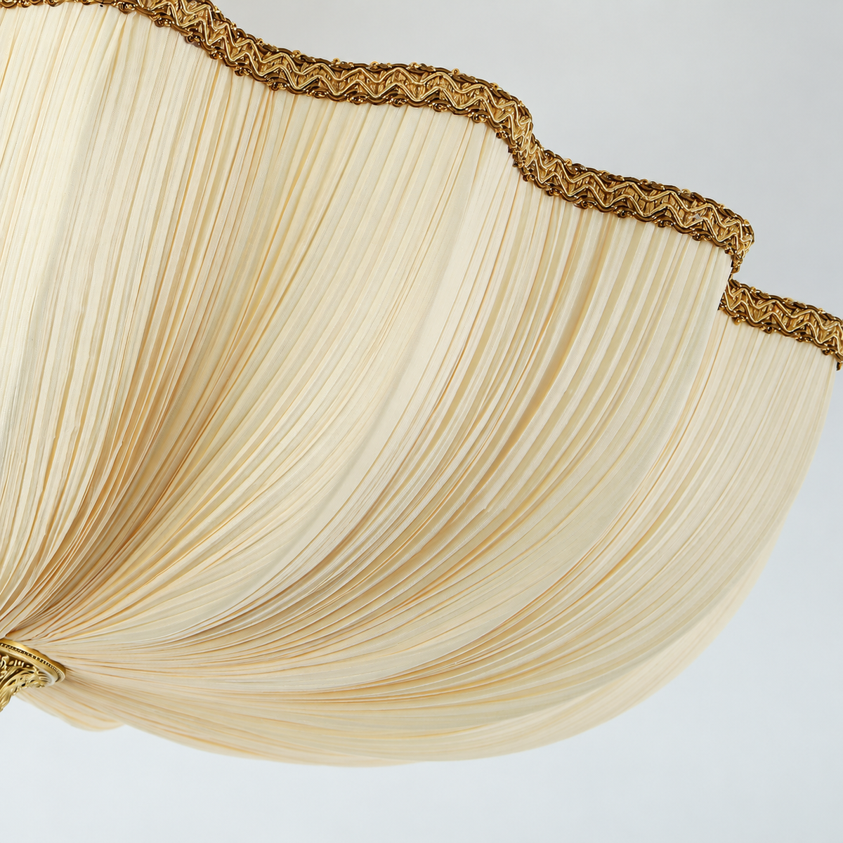 Close-up of a ceiling light with pleated cream fabric and gold-toned braided trim along its scalloped edge against a pale background.