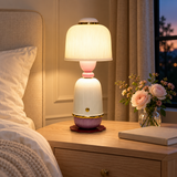 A lit table lamp with a ribbed white shade, pink neck, and purple base sits on a wooden bedside table next to a bed, with flowers and a book nearby in a warm bedroom setting.