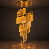 A modern spiral chandelier with clear glass tubes that glow warm yellow when lit, suspended from a circular gold ceiling mount in a dark room.